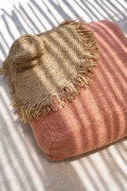 Chapeau large bord raphia naturel tressé frangé sur coussin rose poudré texturé