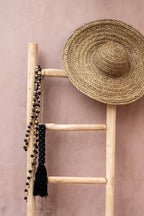 Chapeau paille sombrero avec perles noires et pompon sur échelle boisée contre mur rose