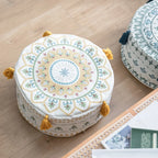 Housse de pouf rond avec motif jaune et pompons, posée sur un sol en bois dans un salon bohème.