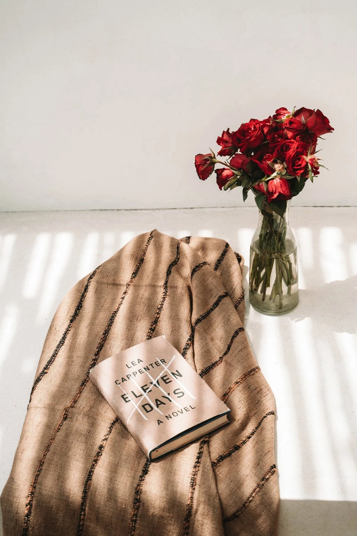 Plaid en coton beige rayé avec franges, livre Eleven Days et vase de roses rouges