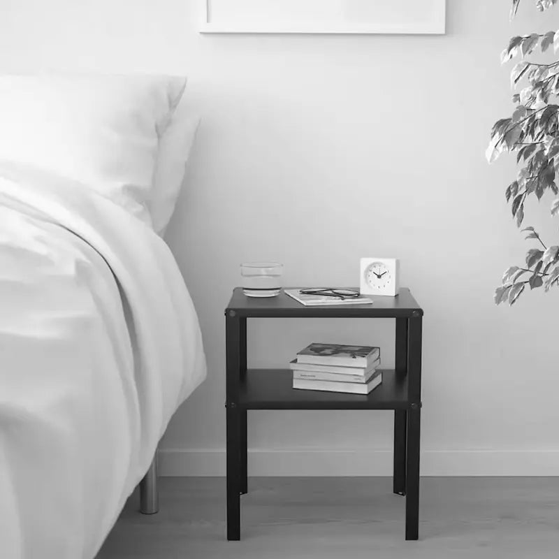 Table de chevet en métal noir, placée près d'un lit dans une chambre à la décoration minimaliste, avec une horloge et des livres.